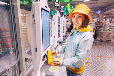 Mutlu genç müşteri kadın, modern bir süpermarkette, kuyrukta beklemekten kaçınmak için bir şişe portakal suyu alır ve parasını öder.