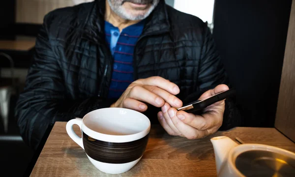 男はカフェで休んでいて お茶を飲んで スマートフォンでメッセージを読んだり書いたりしています モバイルオフィスで働く 一日中のコミュニケーション 被写界深度が浅いクローズアップソフトフォーカス画像 — ストック写真