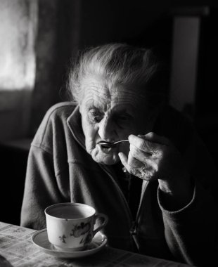 Yaşlı bir depresif kadın siyah beyaz görüntü. Yaşlı bir yalnız kadın mutfağa yakın pencere ve çay içme bir masada oturur. Yandan odaklanmak