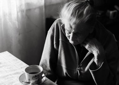 Yaşlı bir depresif kadın siyah beyaz görüntü. Yaşlı bir yalnız kadın mutfağa yakın pencere ve çay içme bir masada oturur.