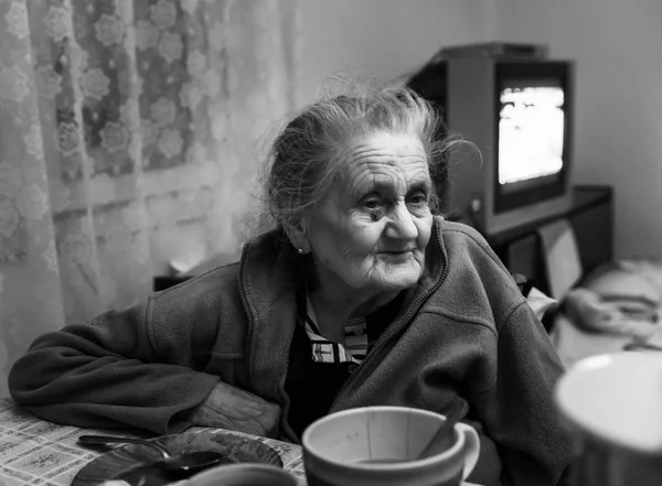 Old depressed woman. Black and white image of an elderly lonely woman sits at a table in the kitchen near the window and drinking tea