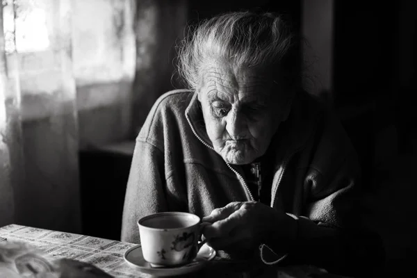 一个沮丧的老太婆的黑白形象 一位孤独的老年妇女坐在窗户附近厨房的一张桌子旁喝茶 — 图库照片