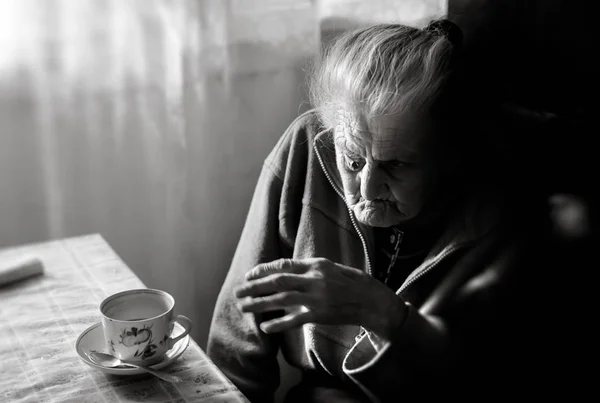 落ち込んでいる老婆の黒と白のイメージ ウィンドウとお茶を飲みながら台所のテーブルに座っている孤独な老女 — ストック写真