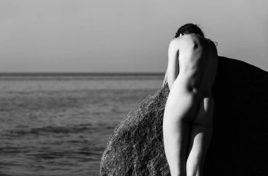 Çıplak kız açık havada doğanın keyfini. Denize karşı büyük taşlar arasında güzel genç çıplak kadın durumda. Siyah beyaz resim