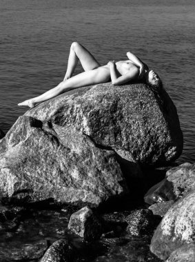 Çıplak kız açık havada doğanın keyfini. Güzel genç çıplak kadın denize karşı büyük taşlar üzerinde yatıyor. Siyah beyaz resim