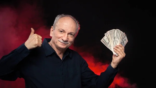拿着一叠钱的老先生 一个兴奋的老商人的肖像 愉快的老人拿着美元钞票在黑暗的红色背景 — 图库照片