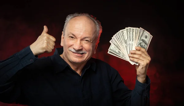 拿着一叠钱的老先生 一个兴奋的老商人的肖像 愉快的老人拿着美元钞票在黑暗的红色背景 — 图库照片