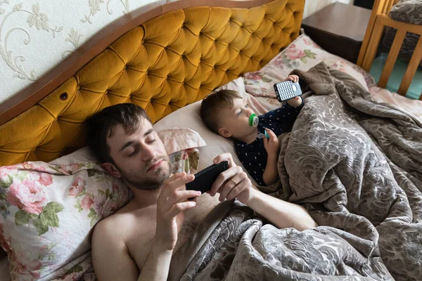 快乐的家庭观念 一个年轻的父亲和他的小儿子躺在床上玩手机 — 图库照片