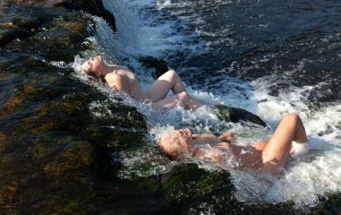 Güzellik, nüdizm ve sağlıklı yaşam tarzı konsepti. Güneşli bir yaz gününde iki çıplak kadın. Genç bir kadın şelalede tazeliğin ve temiz suyun tadını çıkarır.. 