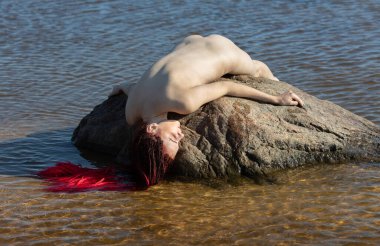 Kızıl saçlı genç bir kadın denizdeki bir taşın üzerinde yatıyor.