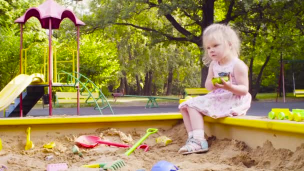 En liten flicka som sitter i en sandlåda är att plocka upp sand — Stockvideo