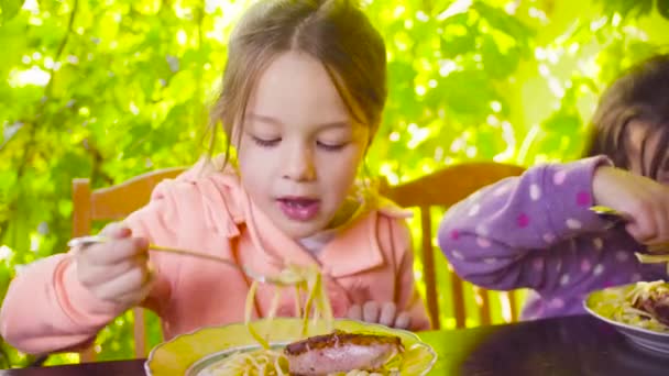 Zwei freche Mädchen sitzen am Tisch und essen Pasta und Bratwurst — Stockvideo