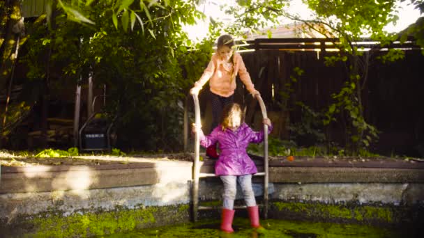 Menina sentada em uma escada de metal e pés pendurados na piscina velha coberto com erva daninha — Vídeo de Stock