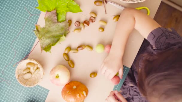 Little girl sitting at the desk makes necklace of acorns — Stock Video