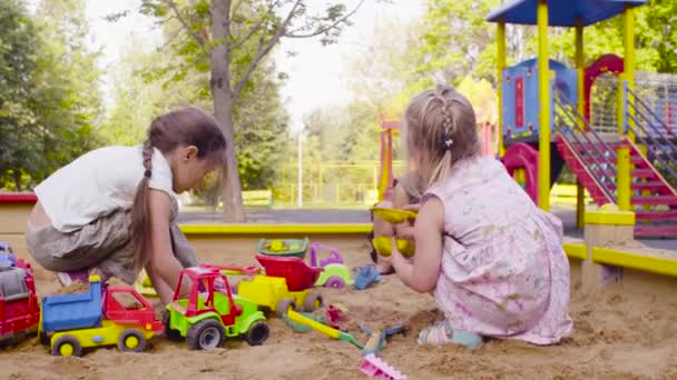 Three girls sitting in a sandbox and picking up sand — Stock Video