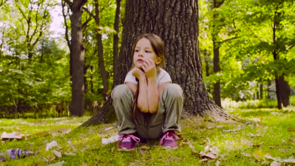 Una chica sentada en el parque en la hierba cerca del árbol — Vídeos de Stock