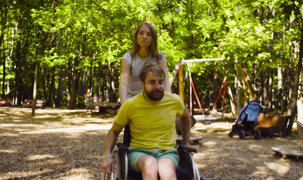 Joven hombre incapacitado en una silla de ruedas caminando en el parque con su esposa —  Fotos de Stock