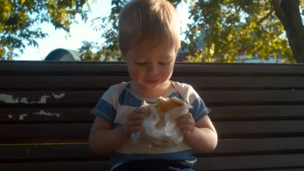Malý chlapec, který sedí na lavici a žere housku — Stock video