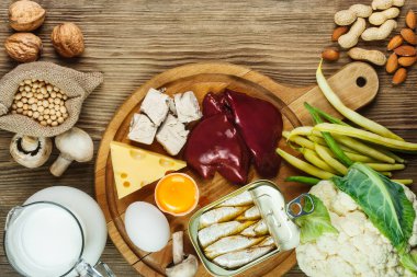 Biotin açısından zengin gıdalar (B7 vitamini). Karaciğer, yumurta sarısı, maya, peynir, sardalya, soya, süt, karnabahar, yeşil fasulye, mantar, fıstık, ceviz ve badem gibi gıdalar ahşap masa üzerinde.