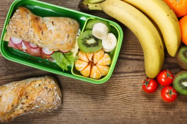 Beslenme çantası ahşap masada. Jambonlu sandviç ve plastik kutuda meyveler.