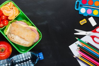 Okul yemeği. Sandviç, küçük domates, mandalina, plastik beslenme çantasında elma ve siyah karatahtanın üzerinde bir şişe su..