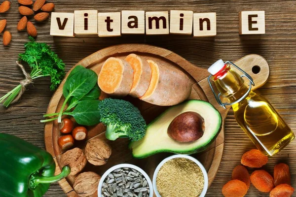 Foods rich in vitamin E such as wheat germ oil, dried wheat germ, dried apricots, hazelnuts, almonds, parsley leaves, avocado, walnuts, sweet potato, broccolii, sunflower seeds, spinach and green paprika on wooden table