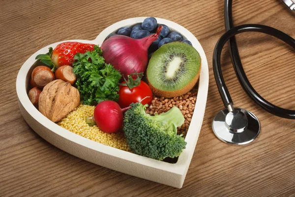 Healthy food in heart shaped bowl and stethoscope. Food such as blueberries, red onion, strawberry, parsley leaves, hazelnuts, walnut, tomato. kiwi, millet, buckwheat, radish, broccoli.