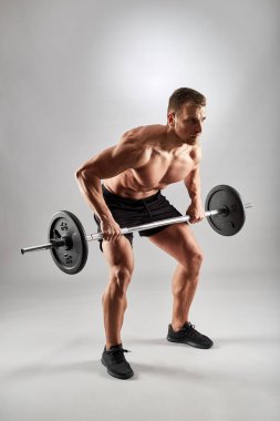 Adam antreman yapıyor, stüdyoda halter sırasını gri arka planda yapıyor.