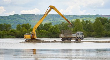 Kazıcı göl kenarındaki bir kamyona kum ve kaldırım taşı yüklüyor.