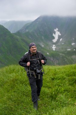 Yağmurluk ve büyük sırt çantalı yürüyüşçü dağlara doğru yürüyor.