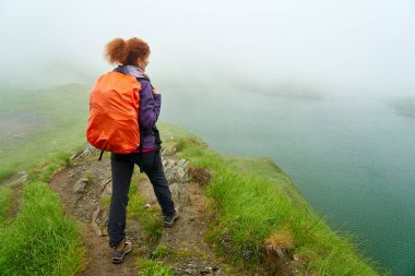 Sırt çantalı bir kadın sislerin arasında buzul gölü kenarında yürüyüş yapıyor.