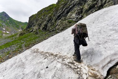 Sırt çantalı bir yürüyüşçü yaz aylarında karda dağ yolunda yürüyüş yapıyor.