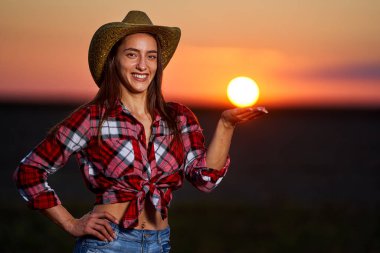 Şapkalı, ekose gömlekli ve kot pantolonlu genç kovboy gün batımında bir tarlada.