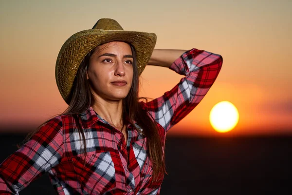 Young cowgirl in hat, plaid shirt and blue jeans shorts at sunset in a field