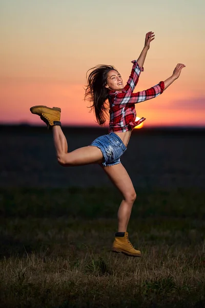 穿着格子呢衬衫和短裤的年轻农妇在日落时分在田里高兴地跳着 — 图库照片