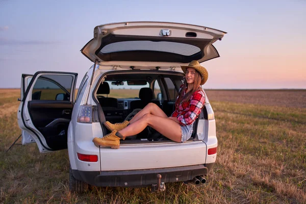 Beautiful young cowgirl watching the sunset sitting in the trunk of her SUV car