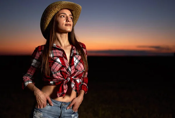 Young cowgirl in hat, plaid shirt and blue jeans shorts at sunset in a field