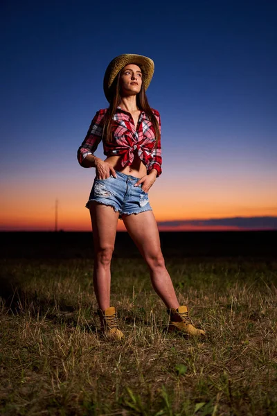 Young cowgirl in hat, plaid shirt and blue jeans shorts at sunset in a field