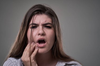Stüdyoda, dişleri ağrıyan, elleri yanağında, gri arka planda genç bir kadın görülüyor.