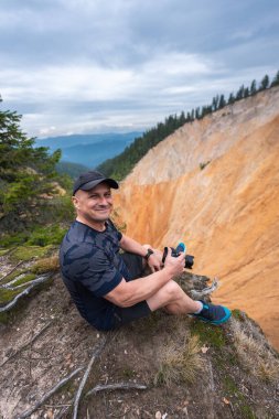 Smiling male hiker holding a camera while sitting on the edge of Groapa Ruginoasa canyon in Apuseni National Park, Romania