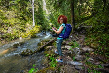 Female hiker with backpack photographing Ponor creek with a smartphone while standing on the rocky forest trail in Apuseni National Park, Romania, a scenic travel destination