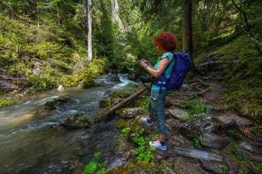Female hiker with backpack photographing Ponor creek with a smartphone while standing on the rocky forest trail in Apuseni National Park, Romania, a scenic travel destination