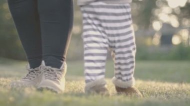Mutlu bebek çocuğun ilk adımları anne yardımı ile çim açık. Düşük açılı yavaş çekim güneşli bir çocukluk aile sahnenin.
