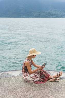 şapka ile kadın cep telefonu kullanarak bir iskelede oturmak