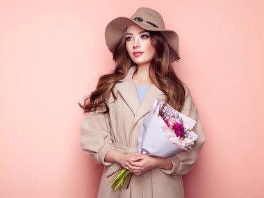 Esmer genç kadın şık Greatcoat ve şapka. Buket çiçek ile bej bir palto kız. Uzun kıvırcık saçlı model. Bahar Lookbook