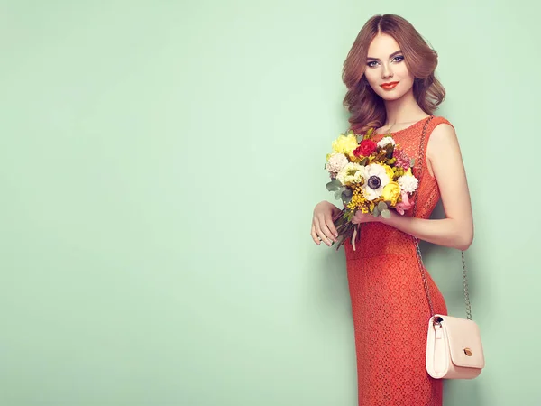 エレガントな赤いドレスで金髪の若い女性 ハンドバッグと緑の背景にポーズの女の子 宝石と髪型 春の花の花束を持つ女性 ファッション写真 — ストック写真