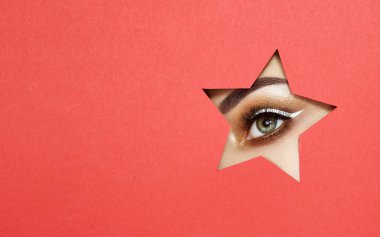 Parlak altın gölgeler ve etkileyici kaşları, genç güzel kadın gözleri yıldız şekilli desen renkli kağıt içine bakar. Yıldız. Noel desenleri. Kırmızı kağıt