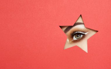The Eyes of the Young Beautiful Woman with Bright Golden Shadows and Expressive Eyebrows, Looks in the Star Shaped Pattern out of Colored Paper. Star. Christmas Patterns. Red Paper clipart