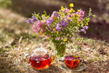 Açık havada çay partisi var. Ormanda bir fincan ve çaydanlığın yakın çekimi. Bir buket çiçek.