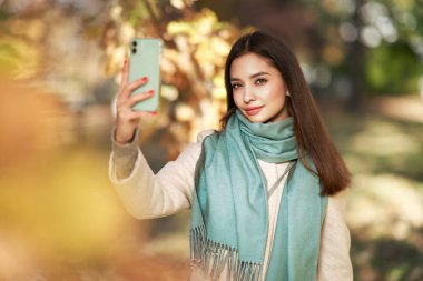 Genç ve güzel bir kadın sonbahar parkında yürüyor. Akıllı telefonlu bir kız sonbahar ormanında fotoğraf çekiyor.